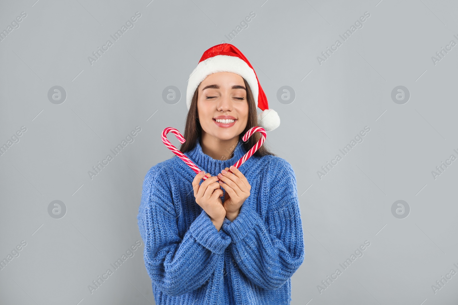 Photo of Young woman in blue sweater and Santa hat holding candy canes on grey background. Celebrating Christmas