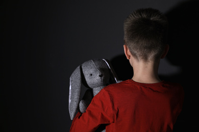 Sad little boy with toy near black wall, back view. Domestic violence concept