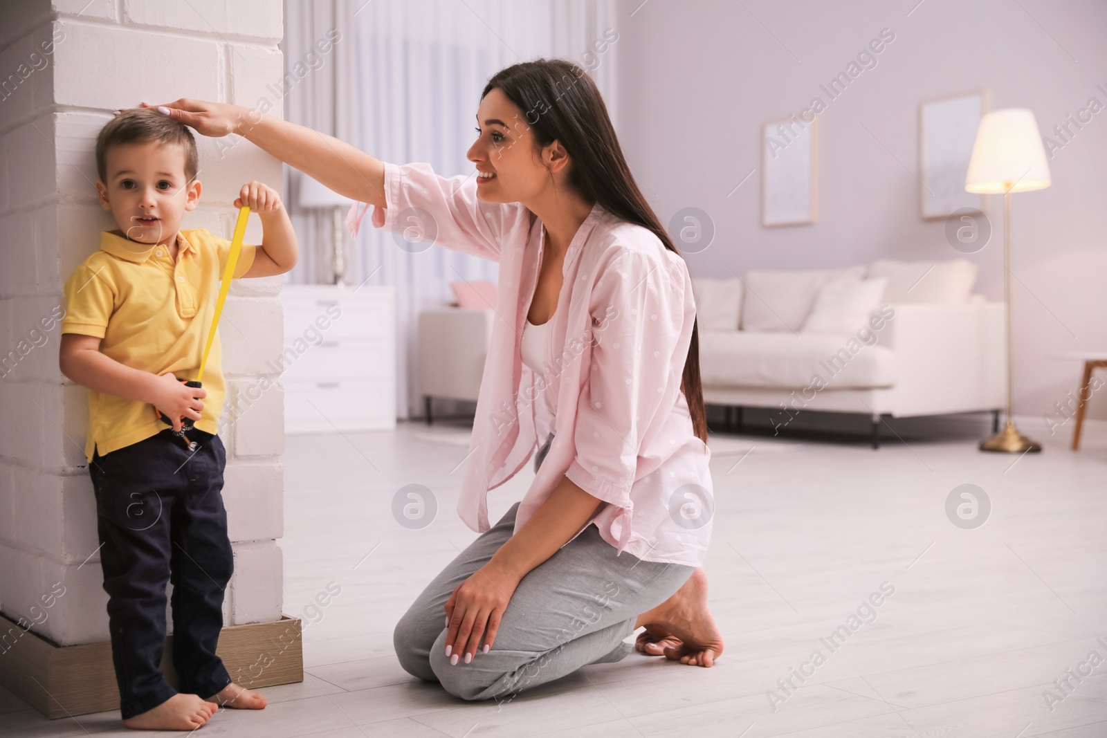 Photo of Mother measuring son's height near white brick pillar at home, space for text