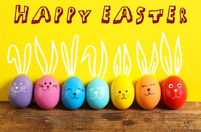 Image of Colorful eggs as Easter bunnies on wooden table against yellow background
