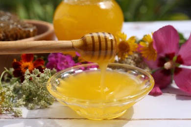 Photo of Delicious honey flowing down from dipper into bowl on white wooden table in garden, closeup