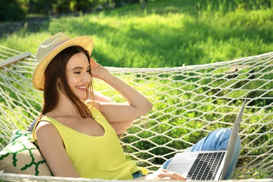 Young woman with laptop resting in comfortable hammock at green garden