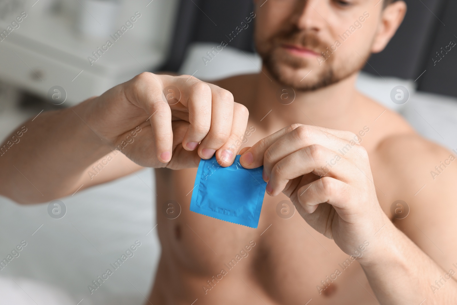 Photo of Closeup view of man opening pack with condom on bed indoors