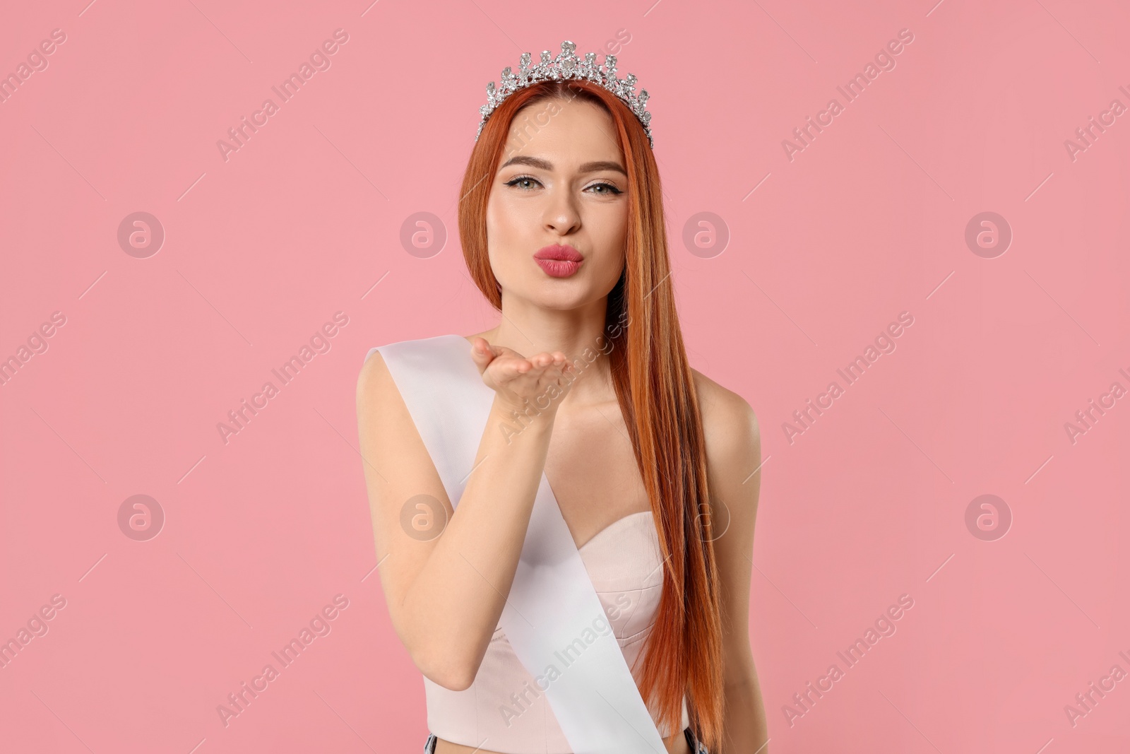 Photo of Beautiful young woman with tiara and ribbon blowing kiss on pink background