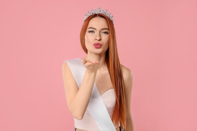 Beautiful young woman with tiara and ribbon blowing kiss on pink background
