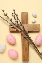 Wooden cross, painted Easter eggs and willow branches on beige background, flat lay