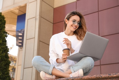 Image of Young woman with paper cup of coffee working on laptop outdoors