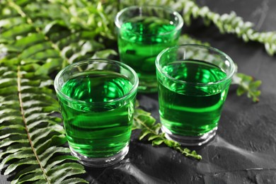 Photo of Absinthe in shot glasses and green leaves on black table, closeup. Alcoholic drink