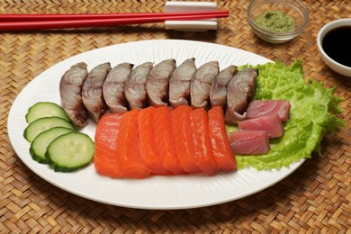 Photo of Delicious mackerel, salmon and tuna served with cucumbers, lettuce, wasabi and soy sauce on wicker mat. Tasty sashimi dish