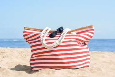Photo of Stylish striped bag with sunglasses on sandy beach near sea
