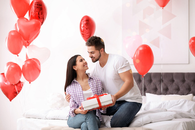 Photo of Young man presenting gift to his girlfriend in bedroom decorated with heart shaped balloons. Valentine's day celebration