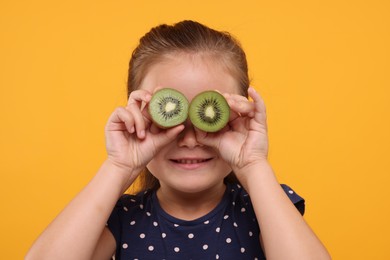 Smiling girl covering eyes with halves of fresh kiwi on orange background