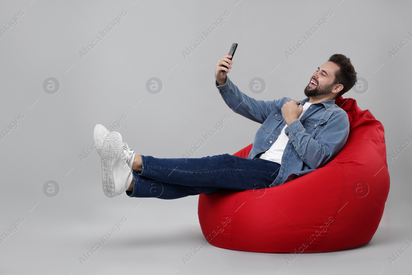 Photo of Happy young man using smartphone on bean bag chair against grey background