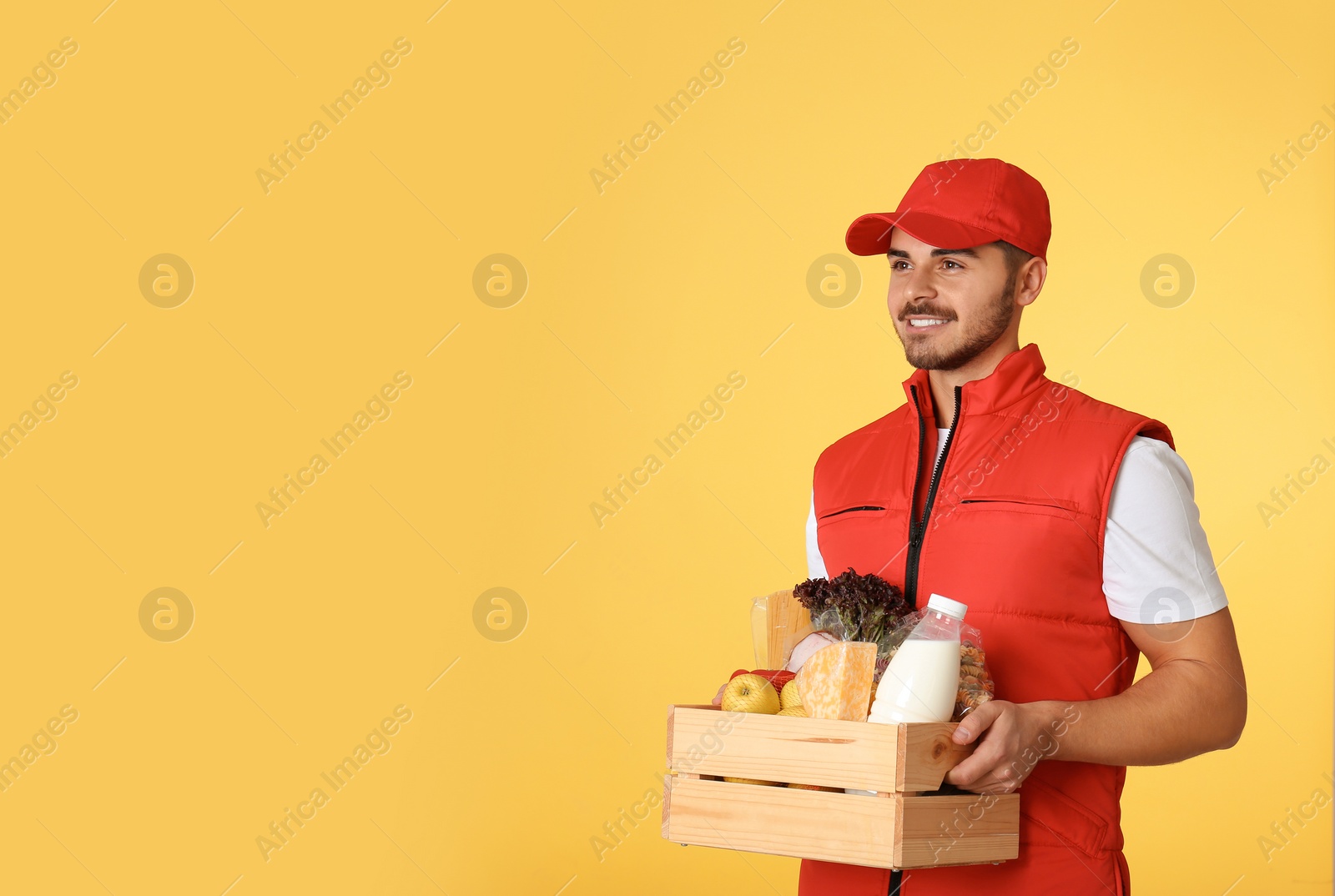 Photo of Food delivery courier holding wooden crate with products on color background. Space for text