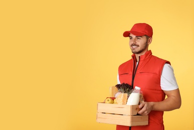 Photo of Food delivery courier holding wooden crate with products on color background. Space for text