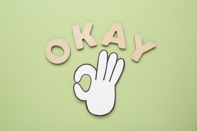 Photo of Word Okay made of wooden letters and paper cutout (OK hand gesture) on pale olive background, flat lay