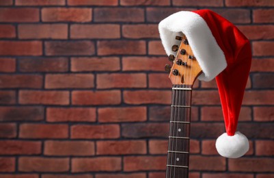 Photo of Guitar with cute Santa hat near brick wall, space for text. Christmas music
