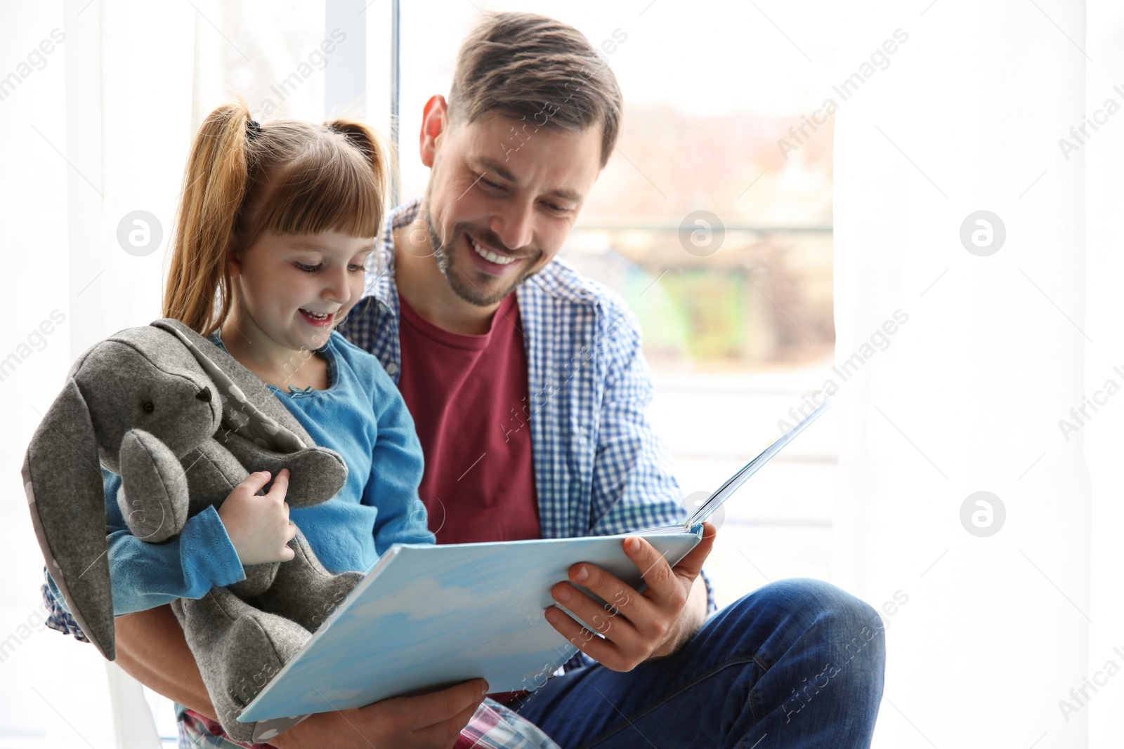 Photo of Happy man and his little daughter reading book near window at home