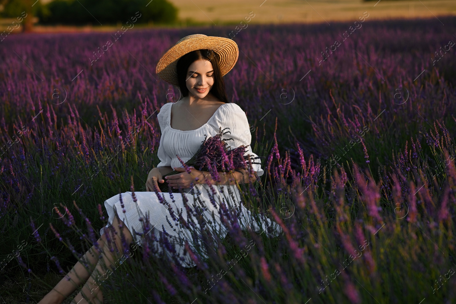 Photo of Beautiful young woman with bouquet sitting in lavender field at sunset