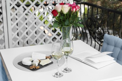 Vase with roses, open book, glasses of wine and snacks on white table at balcony