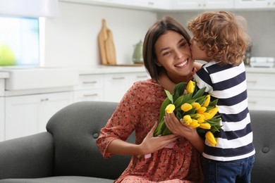 Photo of Little son kissing and congratulating his mom with Mother`s day at home, space for text. Woman holding bouquet of yellow tulips