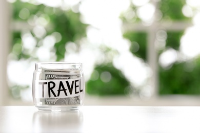 Photo of Glass jar with money and word TRAVEL on table against blurred background, space for text