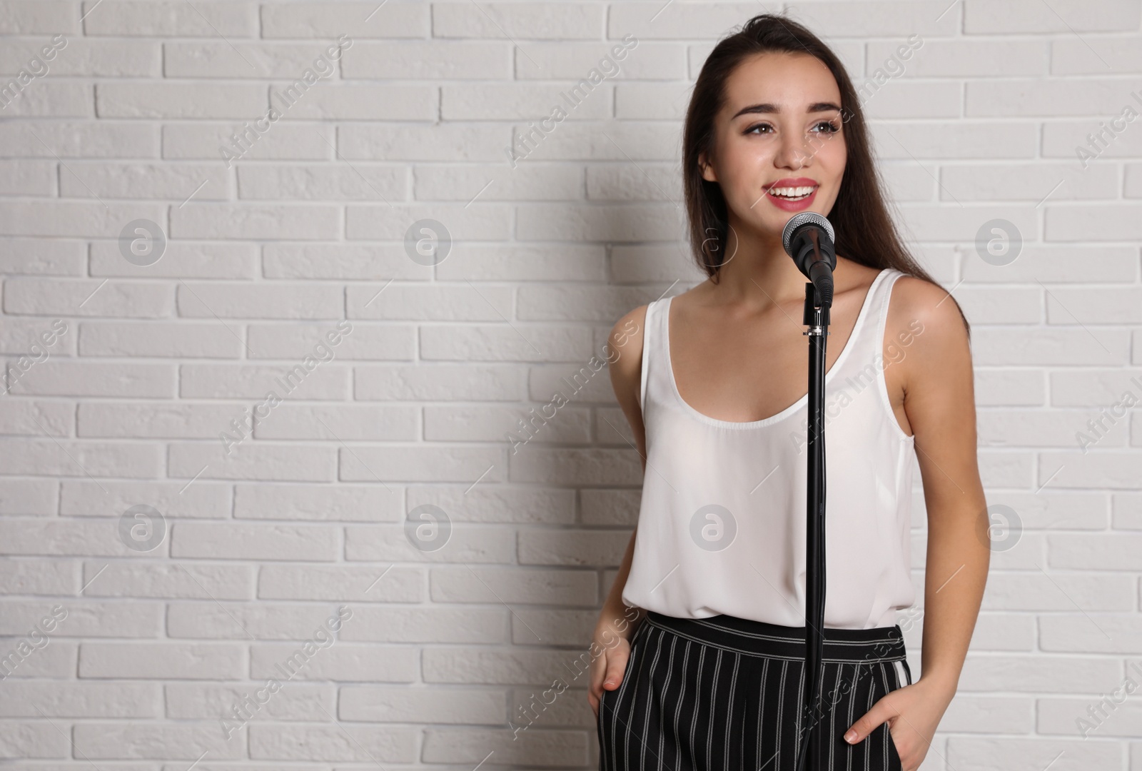 Photo of Young woman in casual clothes posing with microphone near brick wall. Space for text