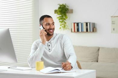 Young man talking on smartphone while working with computer at desk in room. Home office