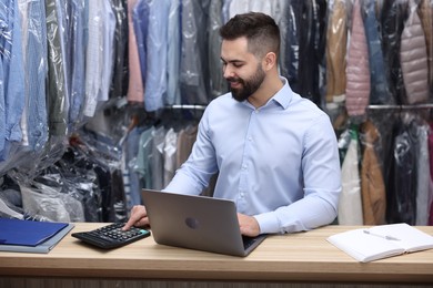 Photo of Dry-cleaning service. Happy worker using calculator while working on laptop at counter indoors