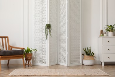 Stylish room with wooden folding screen, armchair and chest of drawers near white wall. Interior design