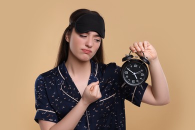 Tired young woman with sleep mask and alarm clock on beige background. Insomnia problem