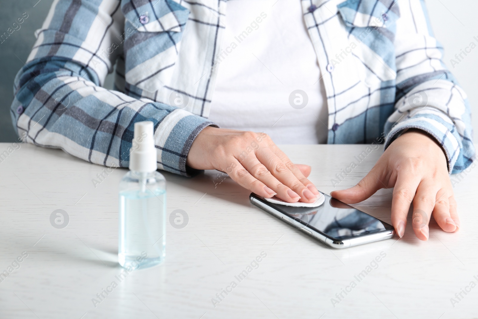 Photo of Woman cleaning mobile phone with cotton pad and antiseptic at white table, closeup