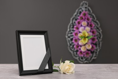Photo frame with black ribbon, rose on light table and wreath of flowers near grey wall indoors, space for text. Funeral attributes