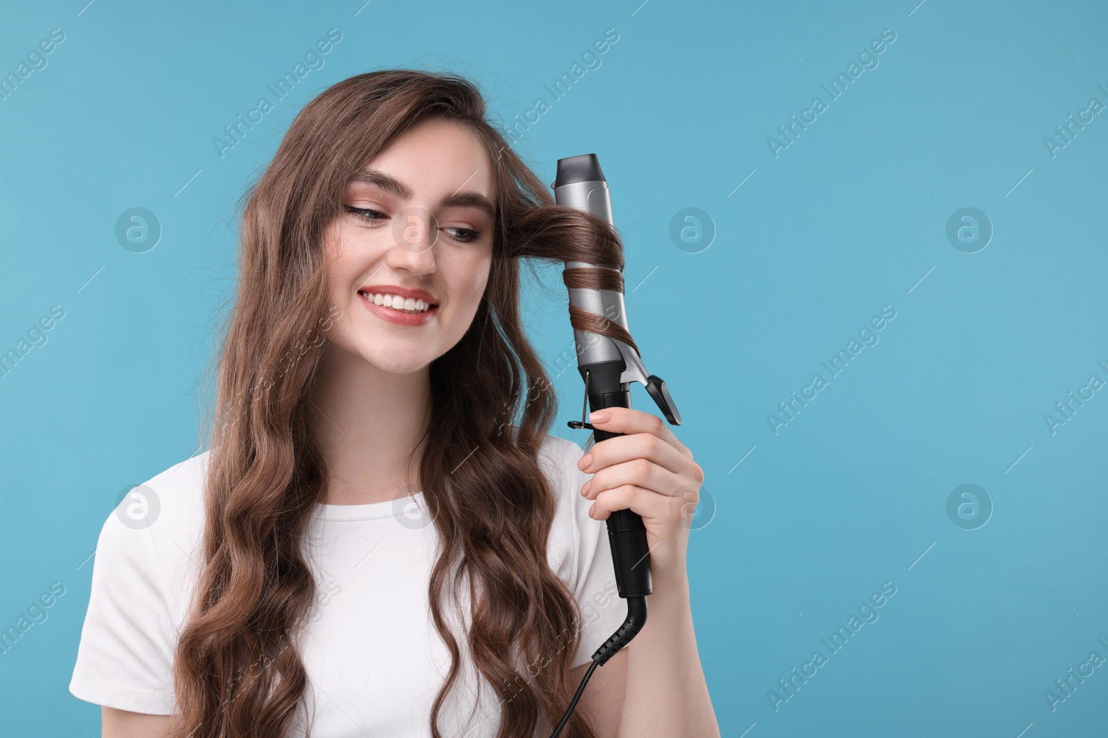 Photo of Beautiful young woman using curling hair iron on light blue background, space for text