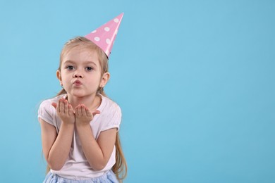 Photo of Birthday celebration. Cute little girl in party hat blowing kiss on light blue background, space for text