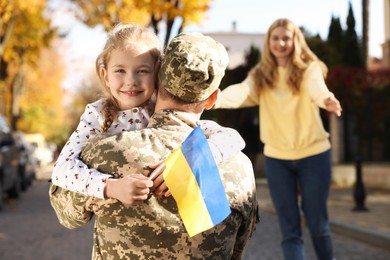 Photo of Daughter hugging her father in military uniform and holding Ukrainian flag on city street. Family reunion