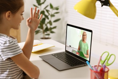 E-learning. Girl raising her hand to answer during online lesson at table indoors