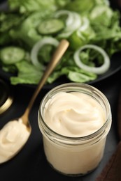 Jar and spoon with delicious mayonnaise near fresh salad on black table