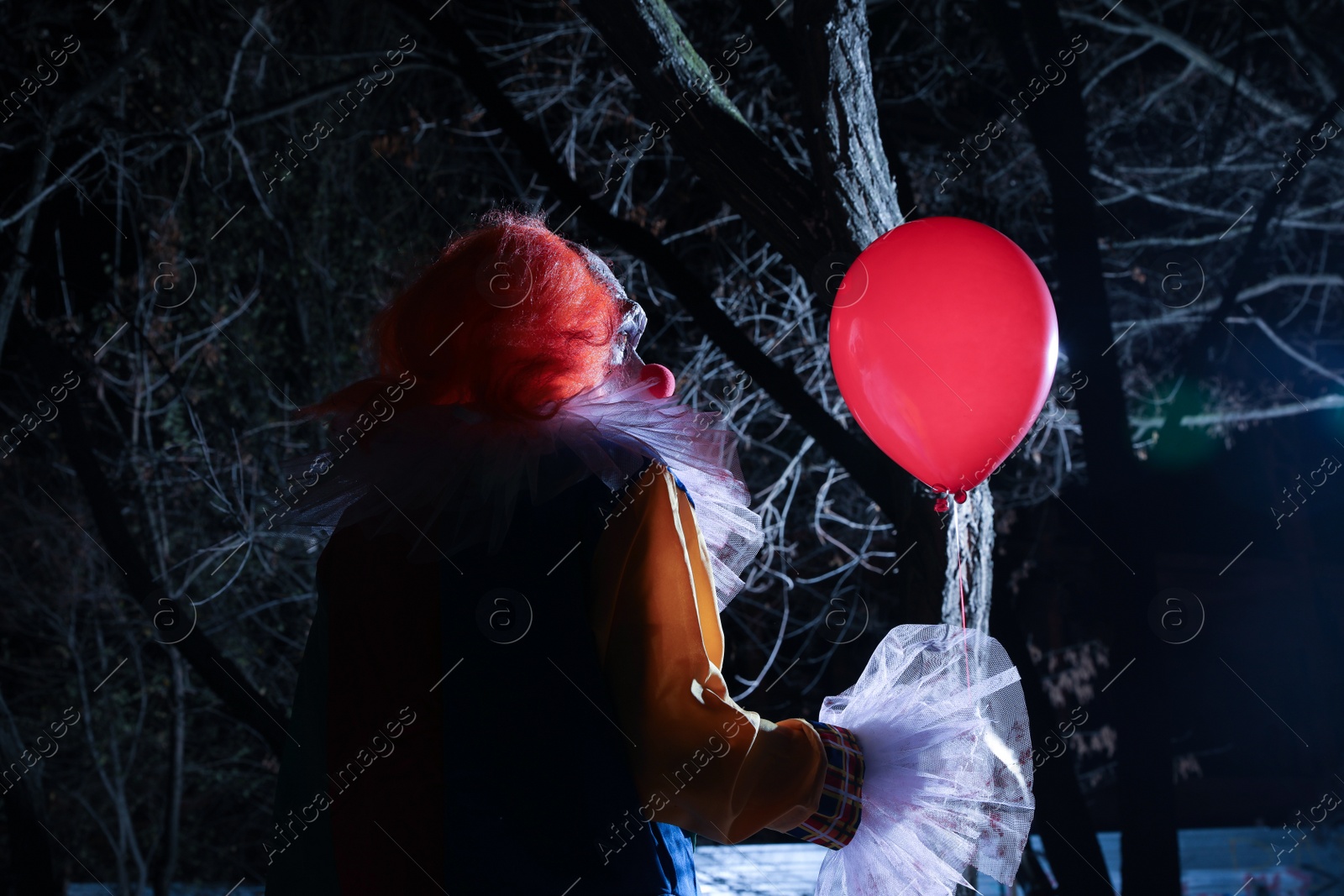 Photo of Terrifying clown with red air balloon outdoors at night. Halloween party costume