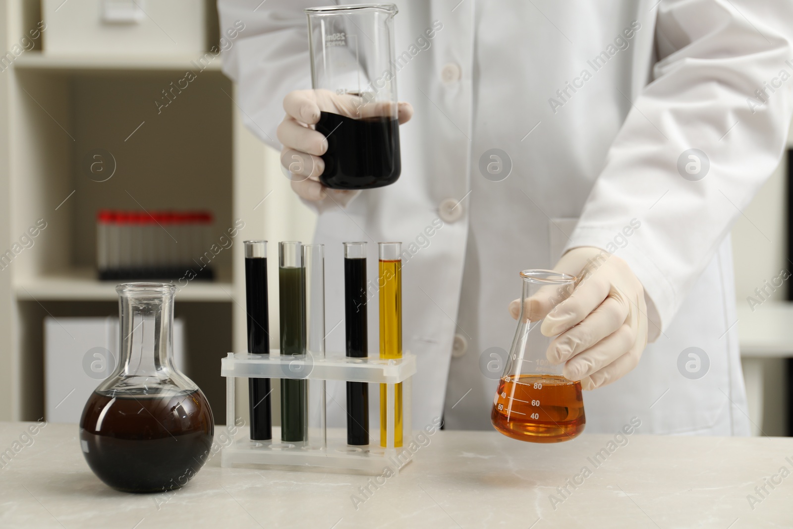 Photo of Laboratory worker holding beaker and flask with different types of crude oil at light marble table, closeup