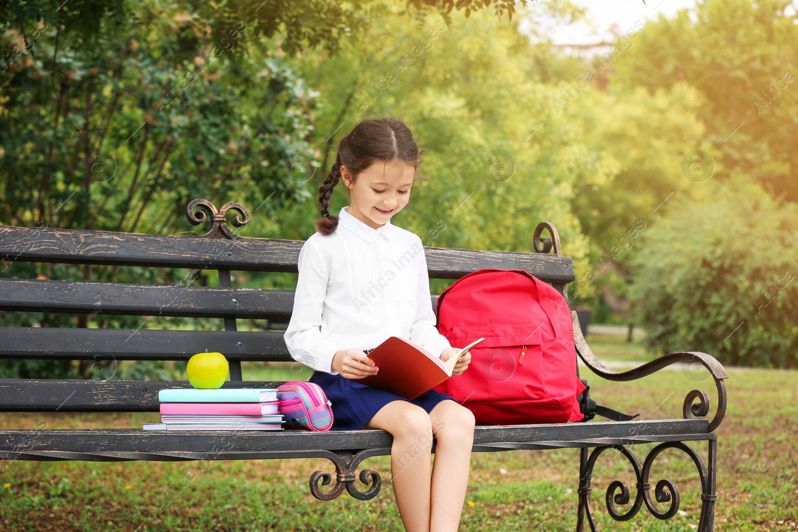 Photo of Cute little school child with stationery reading book on bench in park