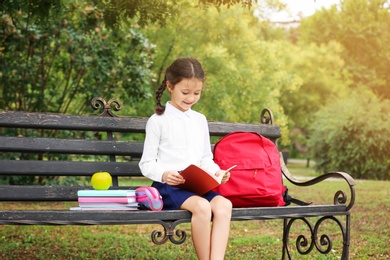 Cute little school child with stationery reading book on bench in park
