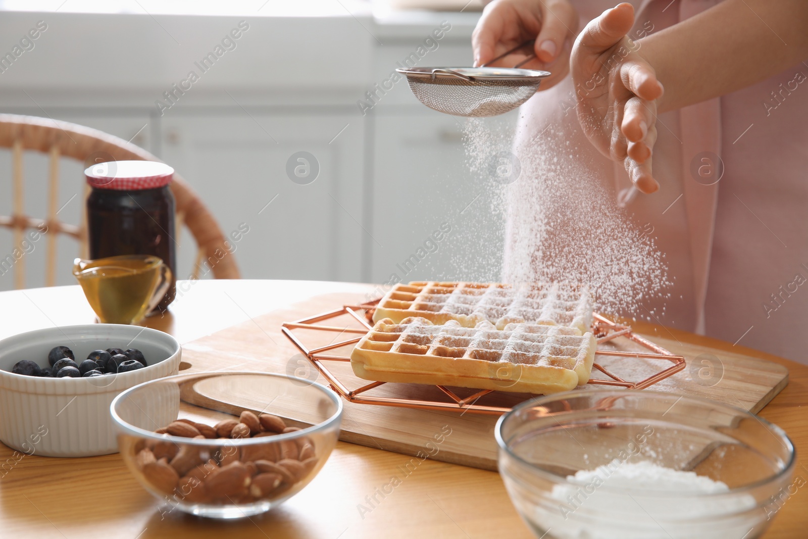 Photo of Woman decorating delicious Belgian waffles with powdered sugar at wooden table in kitchen, closeup