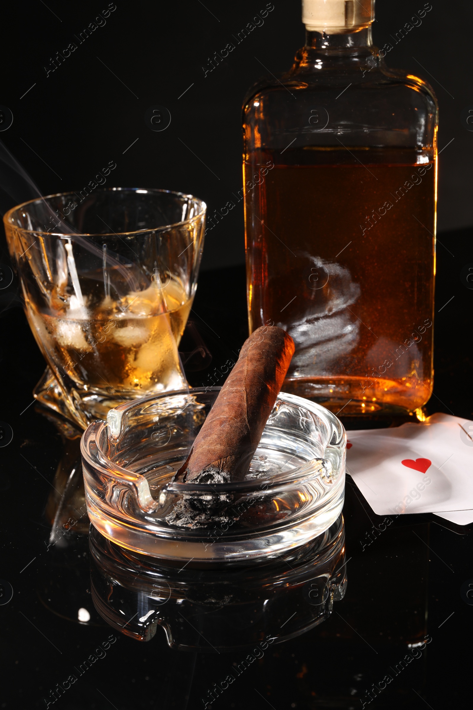 Photo of Smoldering cigar, ashtray, playing cards and whiskey on black mirror surface