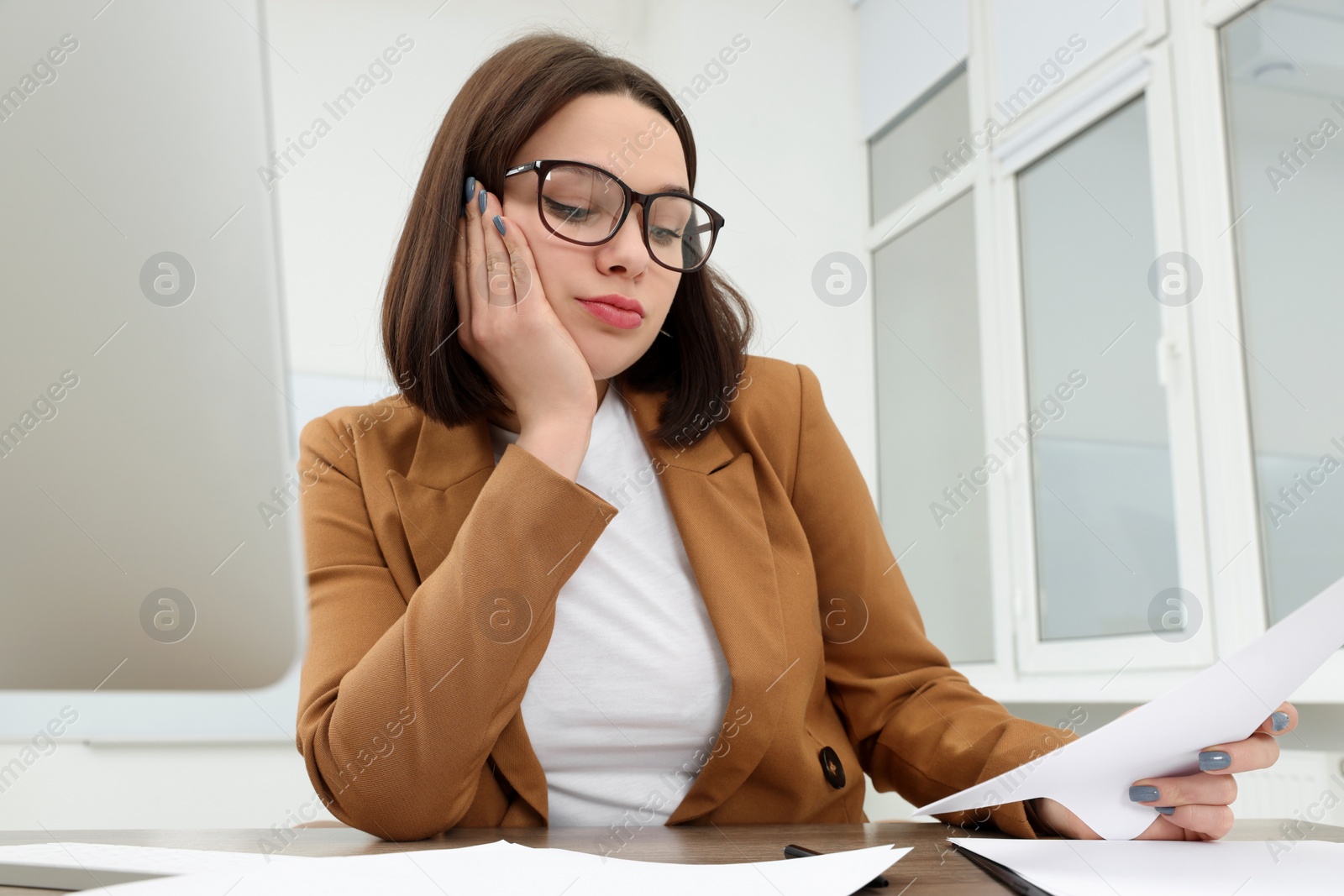 Photo of Puzzled young intern with paper sheets at table in modern office. First work day