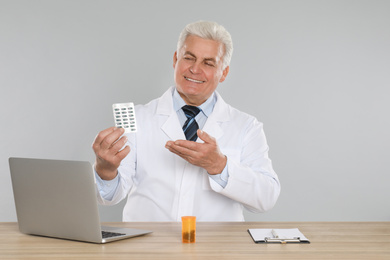 Photo of Professional pharmacist with pills and laptop at table against light grey background