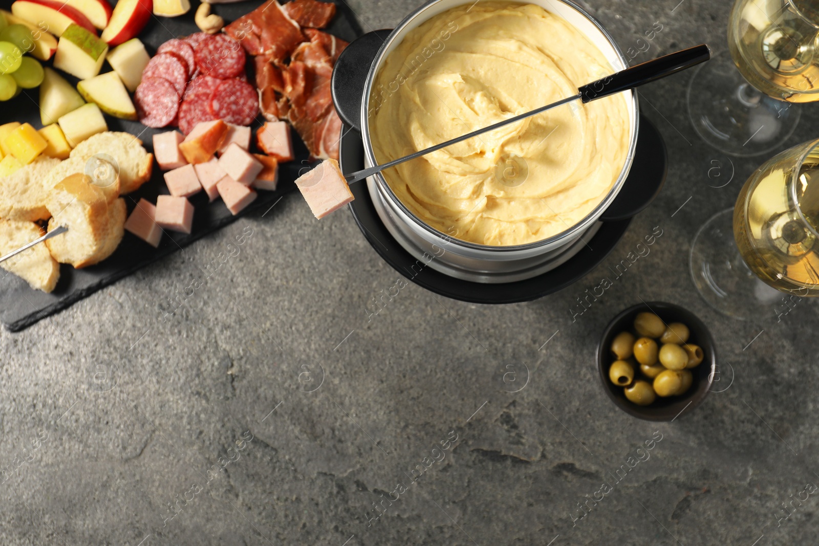 Photo of Fork with piece of ham, fondue pot with melted cheese, wine and snacks on grey textured table, flat lay. Space for text