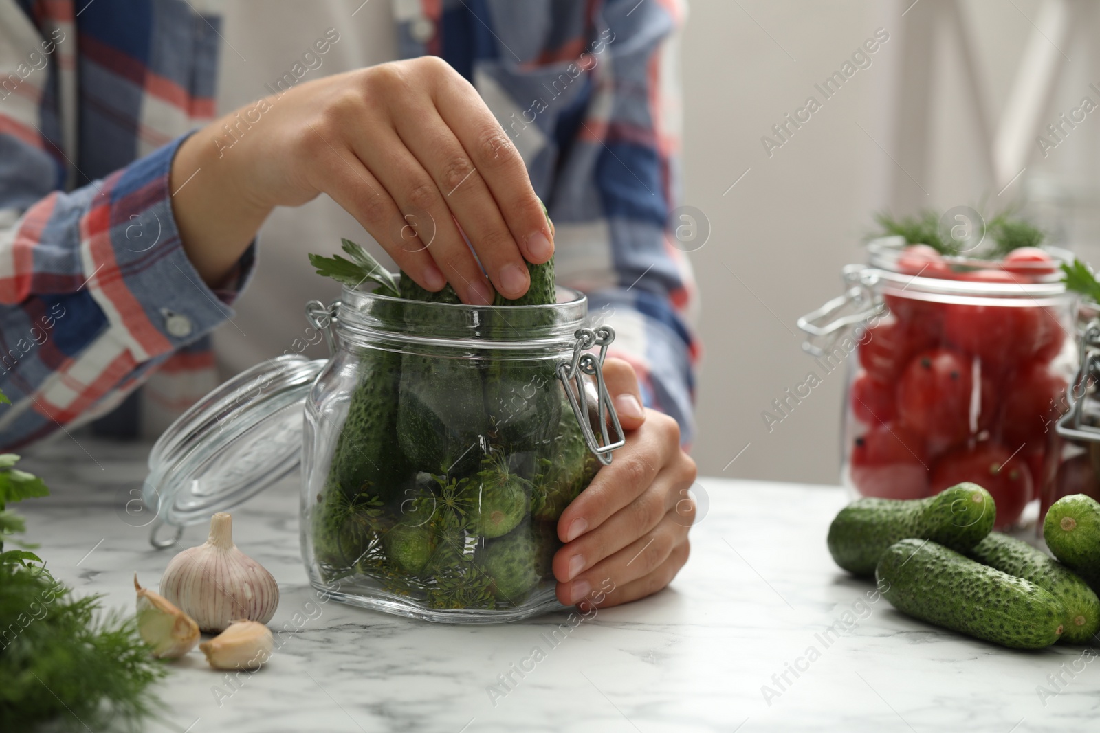 Photo of Woman putting cucumber into glass jar at white marble kitchen table, closeup. Pickling vegetables