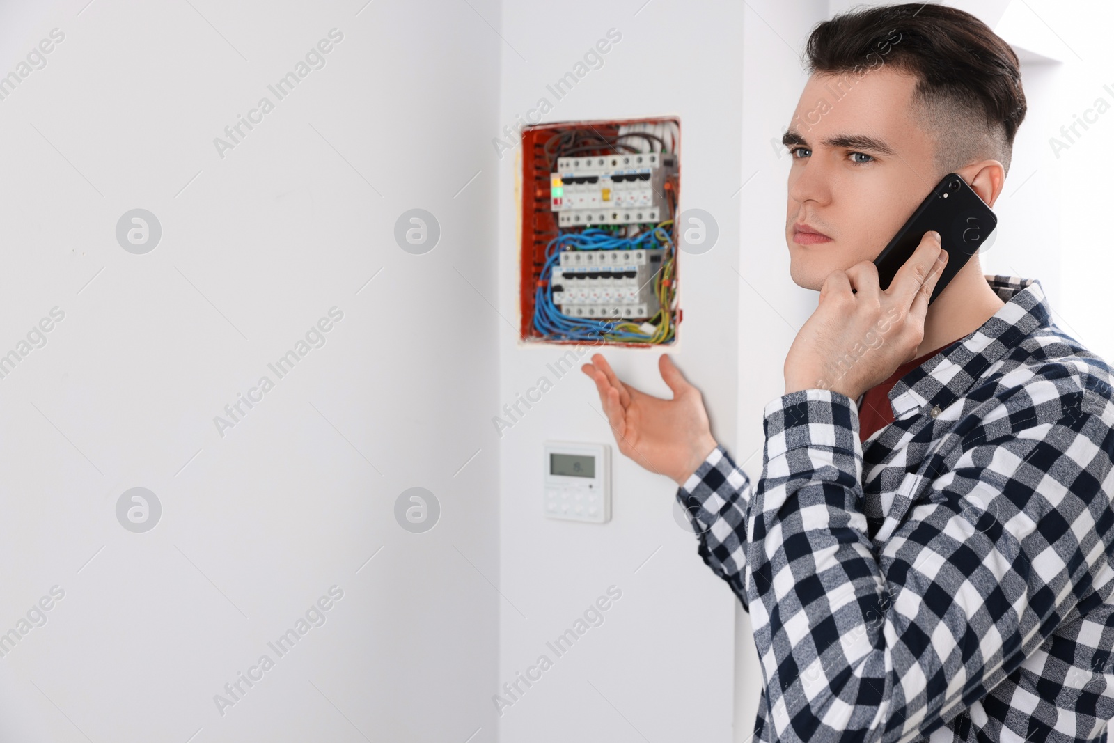 Photo of Young handyman talking on phone near electrical panel board indoors. Space for text