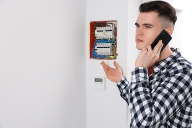 Photo of Young handyman talking on phone near electrical panel board indoors. Space for text